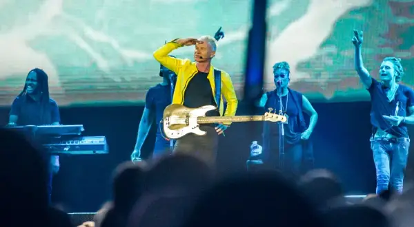 Sting w żółtej kurtce gra na gitarze basowej na scenie z zespołem.