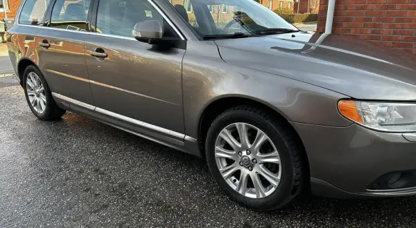 Grå Volvo V70 kombi, en pålitlig bil som kan behöva en ny motorlampa. Bilen står parkerad på en fuktig uppfart.