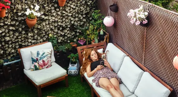 Mujer relajándose en un patio decorado para **decorar patios pequeños con poco dinero**. Flores, luces y muebles crean un oasis acogedor.