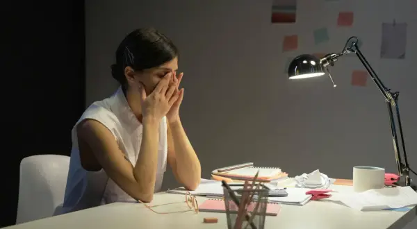 Zmęczona kobieta z rękami na twarzy, siedząca przy biurku po ustaniu zatrudnienia. Lampka biurkowa oświetla stos papierów.