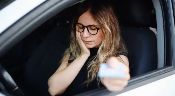 Kobieta w okularach siedzi za kierownicą, trzymając kartę. Wygląda na zmartwioną, jakby czekała na kontrolę, bo grozi jej jazda bez prawa jazdy.