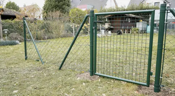 Ein grüner Maschendrahtzaun wird gespannt, mit einem Tor, das offen steht. Im Hintergrund ein Garten mit Häusern und einem Schuppen.