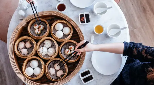 Ręce sięgają po pyszne dim sum z bambusowego parownika. Na stole stoją miseczki z sosem i herbatą.