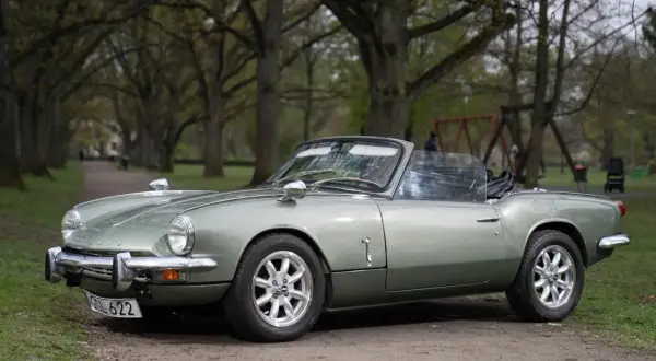 En ljusgrön Triumph Spitfire cabriolet parkerad på en grusväg i en allé.