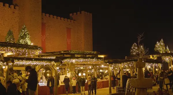 Mercado navideño bajo un castillo iluminado. Gente pasea entre puestos decorados con luces, mostrando como se celebra la navidad.
