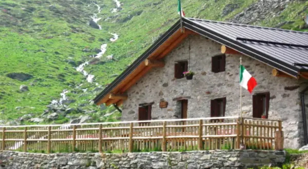 Rifugio in pietra con bandiera italiana, circondato da prati verdi e un ruscello. Un luogo ideale per rifugi val camonica facili.