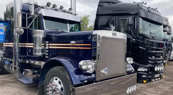 Deux camions américains, un bleu et un noir, brillent sous un ciel couvert. Le bleu, un Peterbilt, arbore des touches orange et beaucoup de chrome.