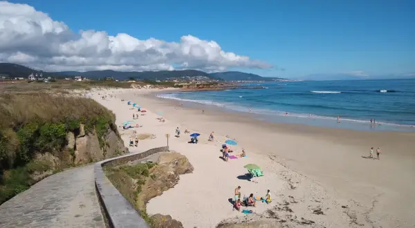 Un paseo de piedra bordea la **playa llas**, donde gente descansa bajo sombrillas y el mar azul invita a un baño.