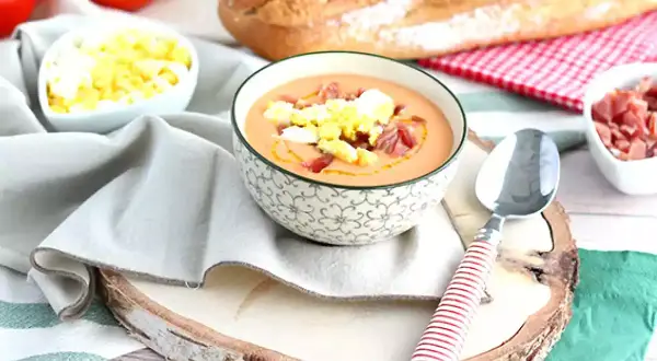 Schüssel mit Gazpacho, garniert mit Ei und Schinken. Daneben sind Tomaten, Brot und weitere Zutaten für kalte Tapas.
