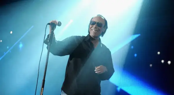 Unico Antonello Venditti sul palco, sorride con occhiali da sole, microfono in mano, illuminato da un fascio di luce blu.