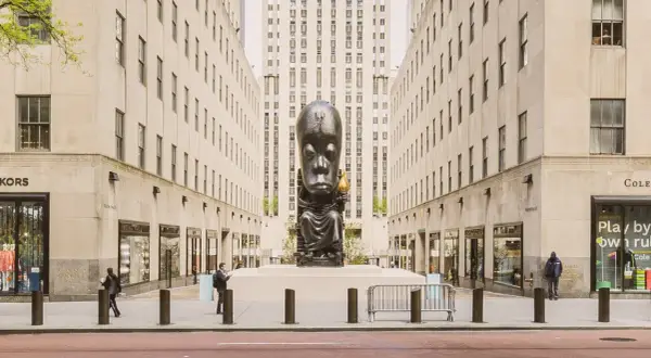 A monumental sculpture of a seated figure with a large head, holding a torch, stands as a striking example of public art between two imposing buildings.