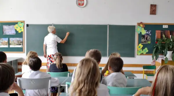 Nauczycielka pisze na tablicy, a uczniowie siedzą w ławkach. To scena z lekcji, która mogłaby się odbywać tuż przed tym, jak skończyło się gimnazjum w Polsce.