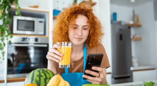 Rudowłosa kobieta z sokiem w dłoni szuka w telefonie informacji, co jeść na poprawę wzroku. Na blacie owoce i warzywa.