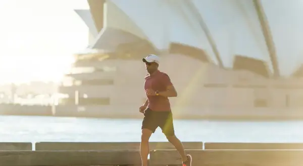 Mężczyzna w czapce i okularach biega przy wodzie, budując bazę tlenową. W tle widać charakterystyczne żagle Opery w Sydney.