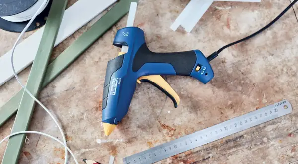 Pistolet à colle bleu et jaune, prêt à l'emploi pour apprendre comment coller du plastique. Des bâtons de colle et des règles sont à proximité.
