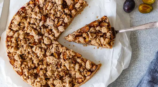 Ein köstlicher, gesunder Pflaumenkuchen mit knuspriger Streuseldecke. Ein Stück wird gerade herausgehoben.