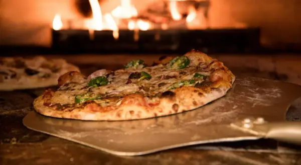 Pizza na łopacie w piecu opalanym drewnem, z płomieniami w tle.