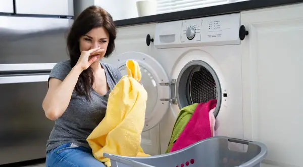 Donna con espressione disgustata annusa un asciugamano giallo, la lavatrice puzza. Cesto con panni colorati accanto.