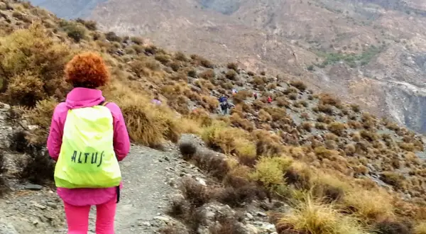 Senderista con mochila amarilla en un sendero montañoso. El grupo avanza, algunos a caballo, en un paisaje árido.