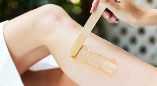 En hand applicerar vax med en träspatel på ett ben för att vaxa bikinilinjen hemma.