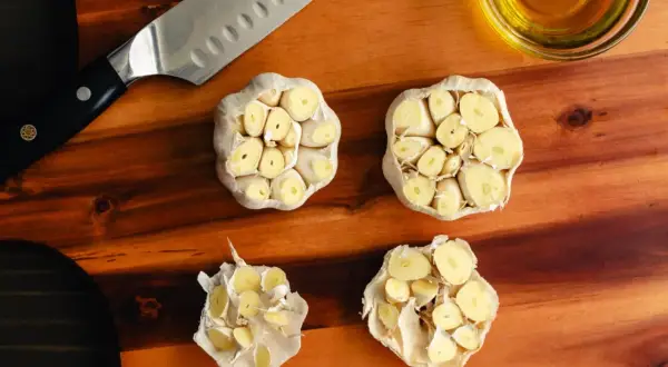 Na desce do krojenia leżą przekrojone główki czosnku, nóż i miseczka z oliwą. Czy czosnek obniża cholesterol?