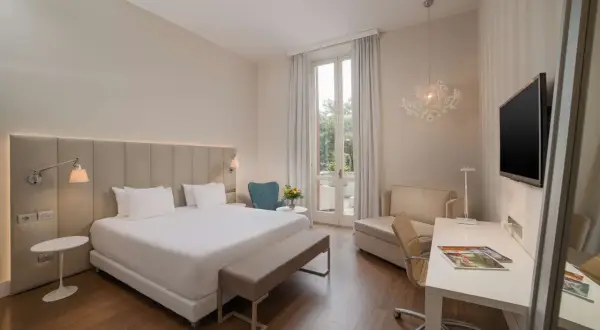 Elegancki pokój hotelowy z dużym łóżkiem, biurkiem i fotelem. Widok na taras z zielenią. Idealne miejsce na wypoczynek w kurorcie na riwierze francuskiej.