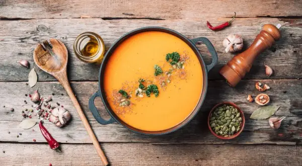 Pomarańczowa zupa dyniowa w garnku, z pestkami dyni, natką i przyprawami na drewnianym stole.