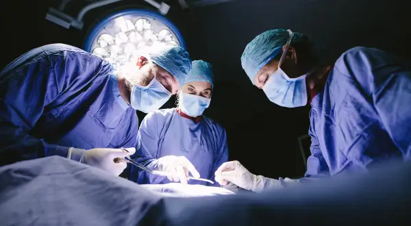 Trois médecins spécialistes en blouse bleue, masqués, concentrés sur une opération sous les lumières vives du bloc opératoire.