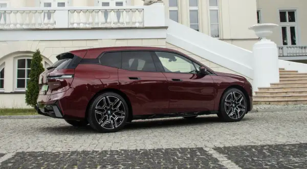 Bordowy BMW iX z eleganckimi felgami stoi przed luksusową posiadłością z kamienną nawierzchnią.