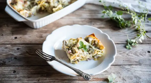 Una fetta di lasagne con carciofi surgelati, guarnita con menta, su un piatto bianco, accanto a una forchetta.