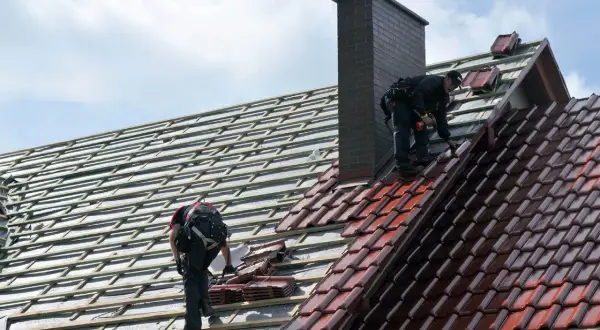 Dwaj dekarze układają dachówki. Widać rusztowanie i komin. Trudno ocenić ile dachówek na m2.