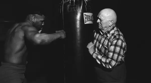 Mike Tyson trains with his manager, a bald man in a plaid shirt, in a dimly lit gym.
