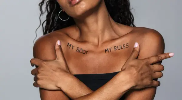 Kobieta z tatuażem na obojczyku "MY BODY MY RULES". Jej ramiona skrzyżowane na piersi, podkreślają siłę i niezależność.