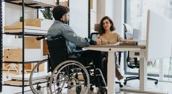 Mężczyzna na wózku inwalidzkim rozmawia z kobietą przy biurku. Pokazuje, że umiarkowany stopień niepełnosprawności a praca w ochronie to nie wykluczające się połączenie.