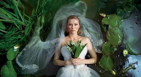 Kirsten Dunst w białej sukni ślubnej leży w wodzie wśród lilii wodnych i trawy. Scena przypomina kadr z jednego z jej filmów.