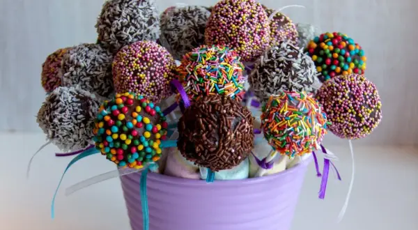 Kolorowe cake pops z jasnego biszkoptu, udekorowane posypkami i wiórkami kokosowymi, w fioletowym wazonie.