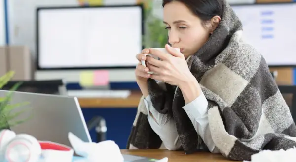 Femme emmitouflée dans une couverture, buvant une tasse de thé, peut-être à cause d'une mauvaise température ambiente.