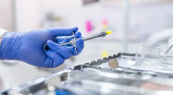 Main d'un dentiste en gant bleu tenant une seringue pour une piqûre aiguille sans saignement. Instruments médicaux en arrière-plan.