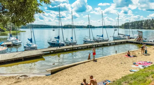 Słoneczny dzień nad jeziorami mazurskimi. Cumujące jachty, piaszczysta plaża z odpoczywającymi ludźmi i błękitne niebo z chmurami.