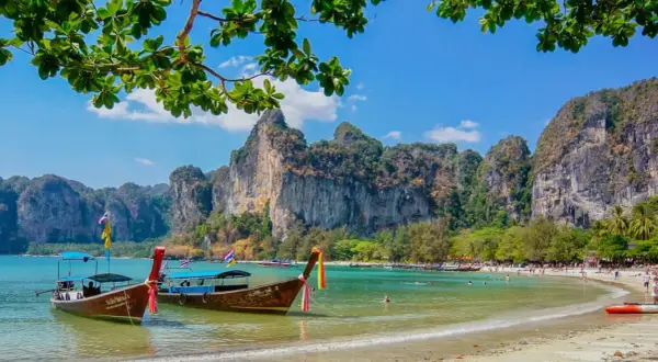 Två longtailbåtar guppar i det turkosa vattnet vid en vacker thailand strand. Imponerande klippor reser sig bakom den gröna djungeln.