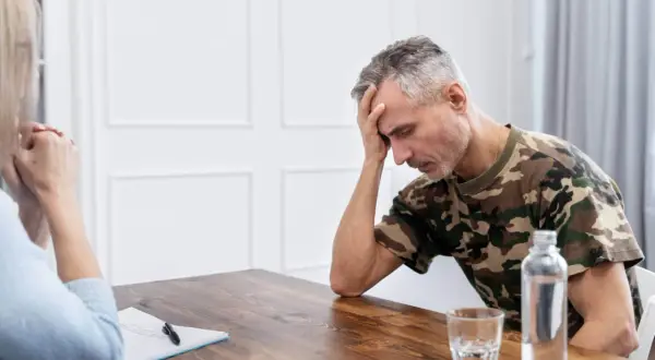 Mężczyzna w mundurze maskującym, z ręką na czole, przeżywa trudne chwile. Rozmowa terapeutyczna, być może przymusowe leczenie alkoholika, jest jego jedyną nadzieją.