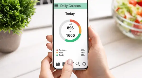 Dłoń trzyma smartfon z aplikacją "Daily Calories", pokazującą spożycie 896 z 1600 kcal. Widoczny jest wzór mifflina w rozkładzie białek, tłuszczów i węglowodanów.