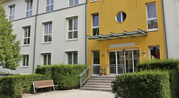 Gelbes Gebäude mit runden Fenster, Eingang mit Glasdach und Stufen. Ein Ort, der §43b SGB XI-Leistungen bietet, lädt zum Verweilen ein.