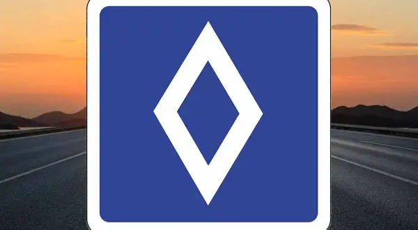 Un nouveau panneau routier bleu avec un losange blanc au centre, sur fond de route et de coucher de soleil.