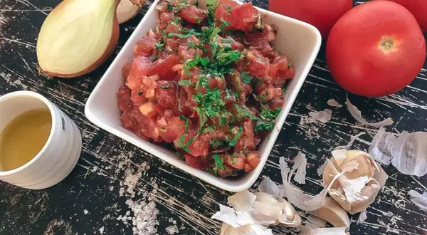 En skål med färsk tomatsalsa, redo för ett tomatsalsa recept. Ingredienser som lök, vitlök och tomater syns runt omkring.