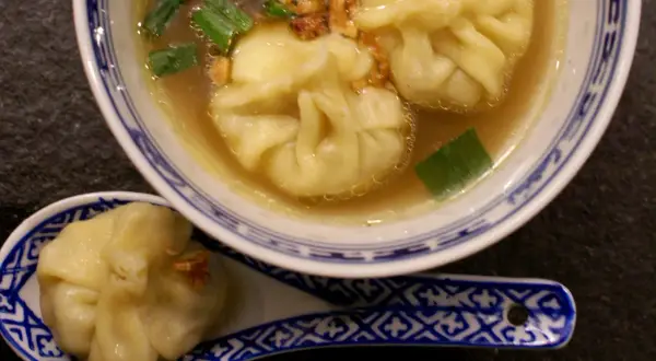 A miseczce z niebiesko-białym wzorem znajduje się aromatyczna zupa wonton z zieloną cebulką i chrupiącymi dodatkami. Na łyżce leży jeden pierożek wonton.