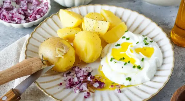 Kartoffeln mit Quark, garniert mit Schnittlauch und verfeinert mit Öl. Dazu gibt es gehackte rote Zwiebeln und ein Messer zum Schälen.