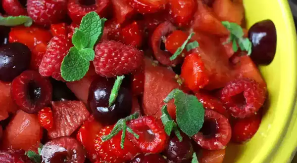 Macedonia di fragole, lamponi e ciliegie, guarnita con foglie di menta fresca, in una ciotola gialla.