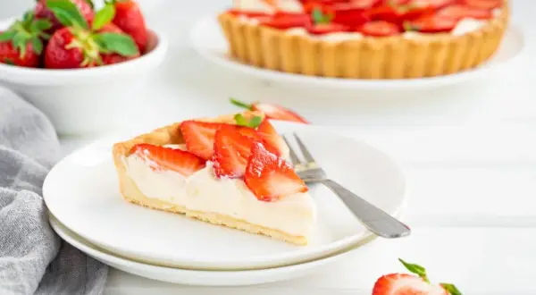 Pyszna tarta truskawkowa z kremowym nadzieniem i świeżymi owocami. Kawałek ciasta na talerzyku, obok cała tarta i miseczka truskawek.