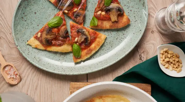 En tunn, krispig flatbread pizza med svamp och basilika, täckt med plastfolie. Pinjenötter och rosa salt ligger bredvid.